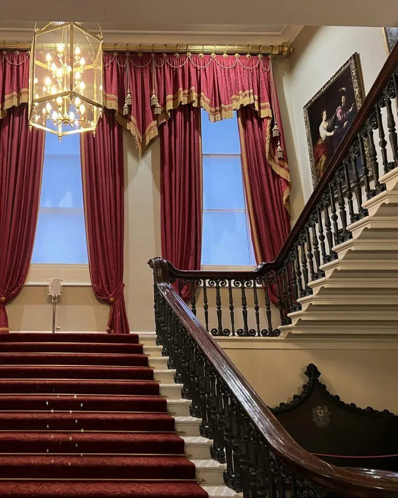 Grand staircase with a red carpet beneath a crystal chandelier and tall draped windows